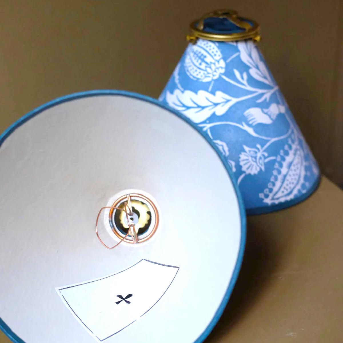 Close-up of a white lampshade with a gold logo and a blue floral-patterned lampshade on a beige surface.